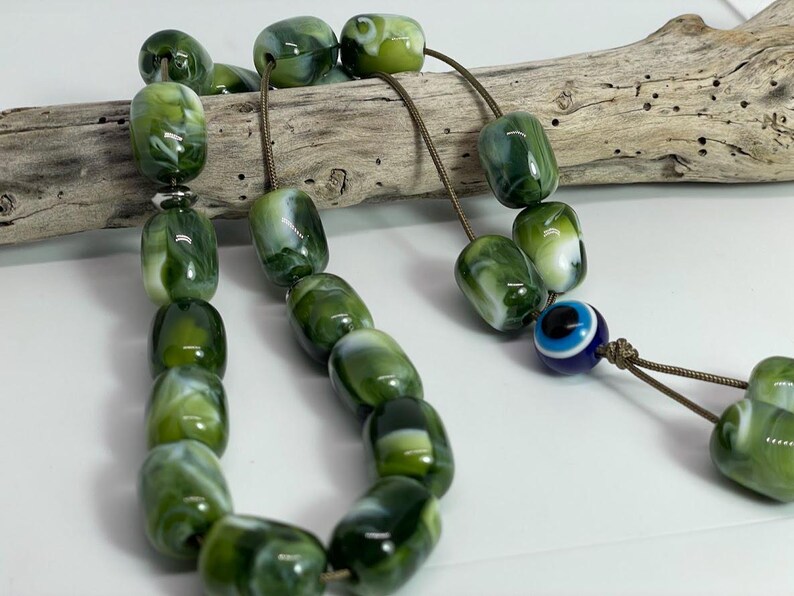 Op de afbeelding: Een groene en witte marmeren gebedskralen ketting met een blauwe kwade oog charme. De kralen zijn geregen op een bruin koord.