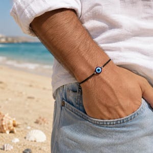 Peut inclure: Un bracelet en cordon noir fin doté d'une breloque centrale circulaire en forme d'œil. La breloque présente une pupille bleu foncé entourée d'un iris bleu clair et d'un anneau blanc, encadrée par deux petites perles métalliques sur le cordon.