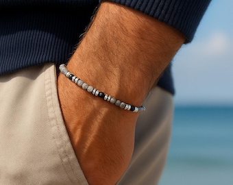 Pode incluir: Uma pulseira de contas com contas cinzentas, pretas e prateadas. A pulseira é usada no pulso, com um suéter azul e calças cáqui no fundo. A pulseira tem aproximadamente 19 cm de circunferência.