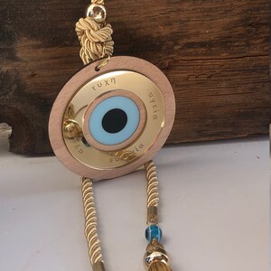 May include: A gold-colored wooden circle with a blue evil eye charm, a gold tassel, and a small gold house charm. The circle has the words "&tau;ύ&chi;&eta;" and "&upsilon;&gamma;&epsilon;ί&alpha;" inscribed on it.