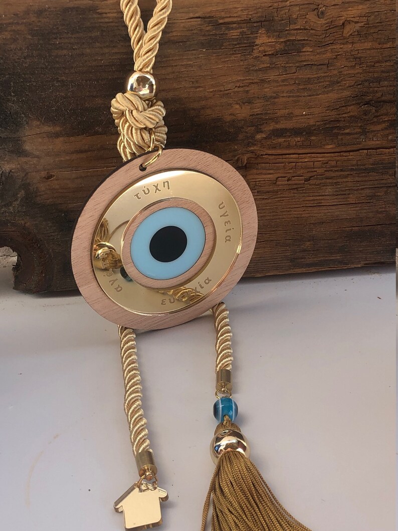 May include: A gold-colored wooden circle with a blue evil eye charm, a gold tassel, and a small gold house charm. The circle has the words "&tau;ύ&chi;&eta;" and "&upsilon;&gamma;&epsilon;ί&alpha;" inscribed on it.