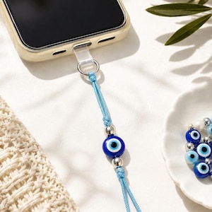Peut inclure: Une breloque de téléphone bleu clair avec une perle centrale en forme d'œil bleu foncé, bleu clair et blanc, encadrée par deux perles métalliques argentées. La breloque est fixée à une languette en plastique transparent insérée dans le port.
