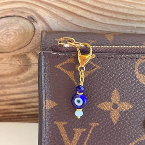 Pu&ograve; includere: Un ciondolo color oro con una perlina a occhio blu e perline blu chiaro e blu scuro. Il ciondolo &egrave; attaccato a un fermaglio e una catena color oro. Il ciondolo &egrave; appeso a un portafoglio marrone con cerniera.
