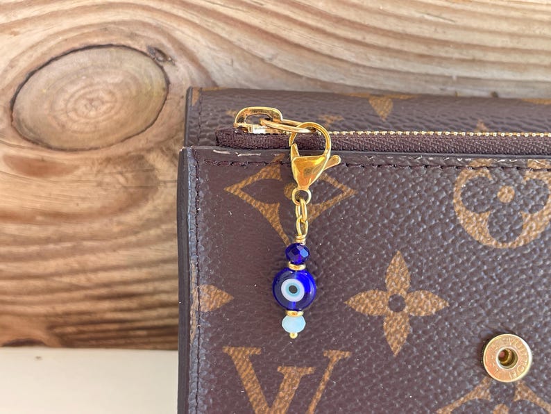 Pu&ograve; includere: Un ciondolo color oro con una perlina a occhio blu e perline blu chiaro e blu scuro. Il ciondolo &egrave; attaccato a un fermaglio e una catena color oro. Il ciondolo &egrave; appeso a un portafoglio marrone con cerniera.