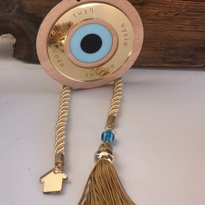 May include: A gold-colored wooden circle with a blue evil eye charm and the words "&tau;ύ&chi;&eta;", "&upsilon;&gamma;&epsilon;ί&alpha;", and "&epsilon;&upsilon;&tau;&upsilon;&chi;ί&alpha;" inscribed around the eye. The circle is attached to a gold rope with a tassel and a small gold house charm.