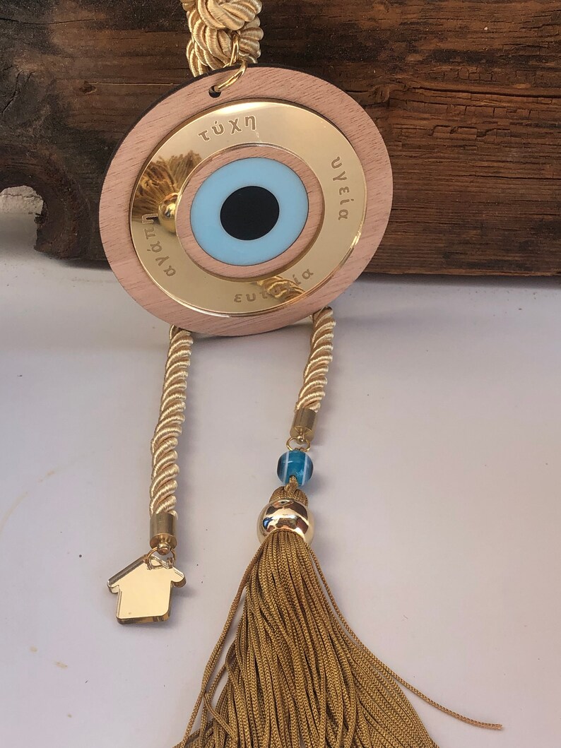 May include: A gold-colored wooden circle with a blue evil eye charm and the words "&tau;ύ&chi;&eta;", "&upsilon;&gamma;&epsilon;ί&alpha;", and "&epsilon;&upsilon;&tau;&upsilon;&chi;ί&alpha;" inscribed around the eye. The circle is attached to a gold rope with a tassel and a small gold house charm.