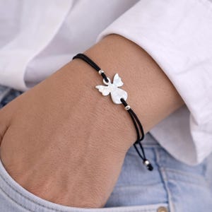 Puede incluir: Pulsera de cordón negro con un dije de ángel plateado. El dije de ángel tiene un diseño de alas detallado y está conectado al cordón con pequeñas cuentas plateadas. La pulsera se usa en la muñeca, sobre un fondo de camisa blanca y vaqueros azules.