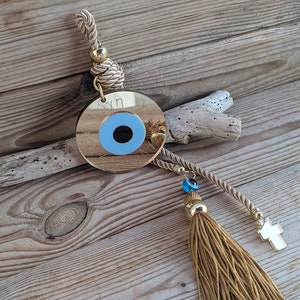 Op de afbeelding: Een gouden boze oog-hanger met een blauw oog en een kwast die aan een touw hangt. De hanger hangt aan een houten oppervlak.