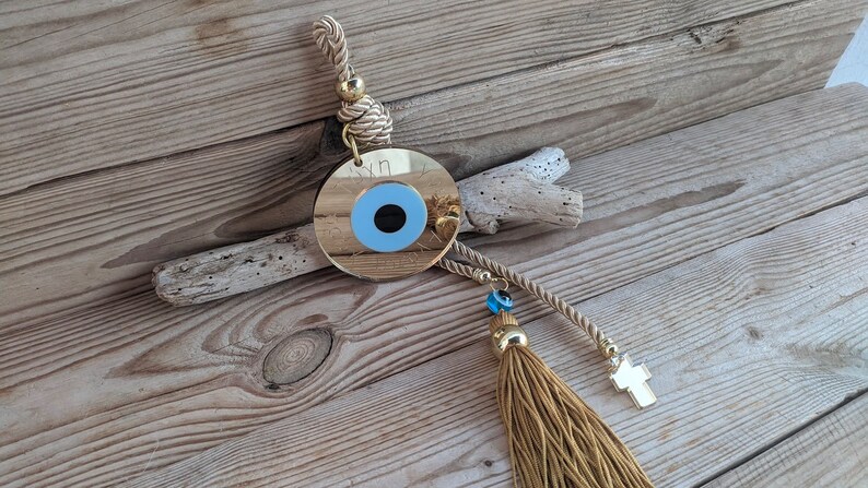 Op de afbeelding: Een gouden boze oog-hanger met een blauw oog en een kwast die aan een touw hangt. De hanger hangt aan een houten oppervlak.