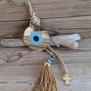 Op de afbeelding: Gouden bedel met een blauw boze oog en een gouden kwast. De bedel is bevestigd aan een gouden touw met een klein gouden kruis aan het einde.