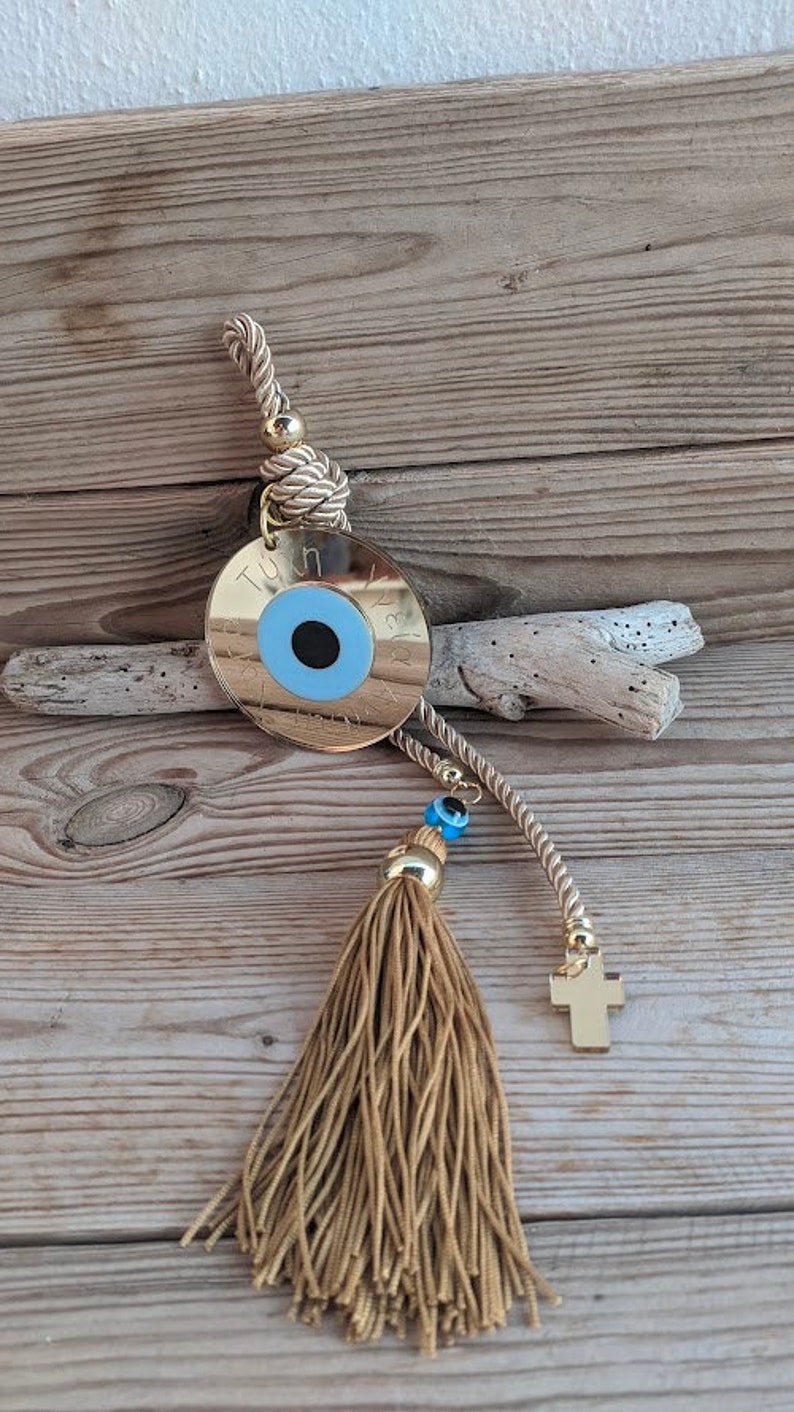 Op de afbeelding: Gouden bedel met een blauw boze oog en een gouden kwast. De bedel is bevestigd aan een gouden touw met een klein gouden kruis aan het einde.