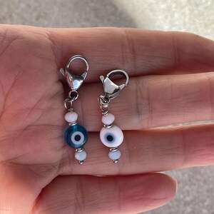 Pu&ograve; includere: Due ciondoli di perline con chiusure color argento. Un ciondolo presenta un design a occhio blu, l'altro un design a occhio bianco. Entrambi i ciondoli hanno piccole perline bianche e azzurre. I ciondoli sono probabilmente per gioielli o accessori.