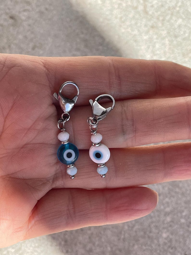 Pu&ograve; includere: Due ciondoli di perline con chiusure color argento. Un ciondolo presenta un design a occhio blu, l'altro un design a occhio bianco. Entrambi i ciondoli hanno piccole perline bianche e azzurre. I ciondoli sono probabilmente per gioielli o accessori.