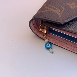 Pu&ograve; includere: Un portafoglio marrone e rosa con un ciondolo color oro. Il ciondolo presenta una perlina blu a forma di occhio, perline bianche e una chiusura color oro. Il portafoglio ha un motivo monogramma.