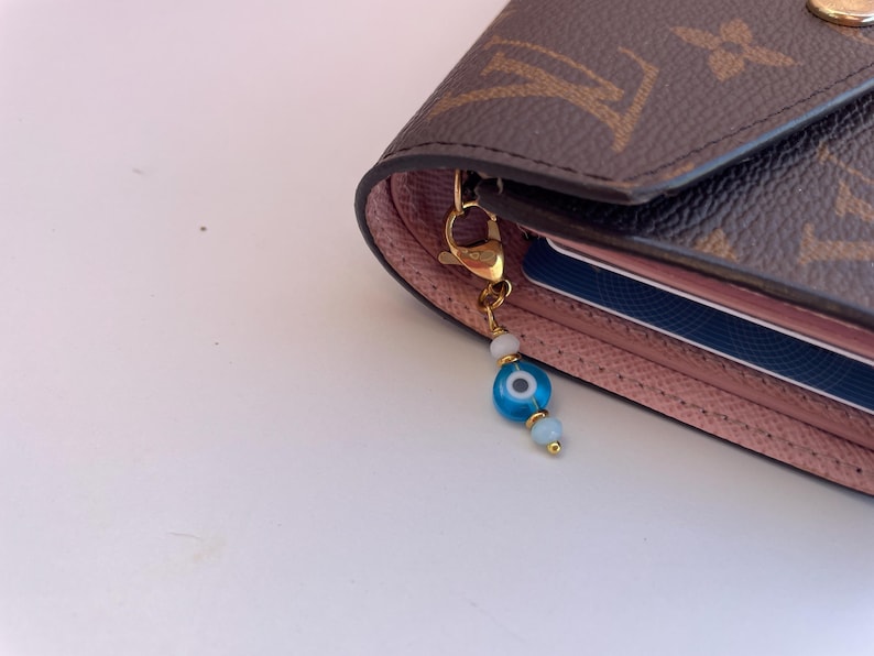 Pu&ograve; includere: Un portafoglio marrone e rosa con un ciondolo color oro. Il ciondolo presenta una perlina blu a forma di occhio, perline bianche e una chiusura color oro. Il portafoglio ha un motivo monogramma.