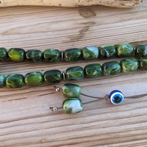 Op de afbeelding: Een groene en witte gebedskralenketting met een blauwe kwade oog charme. De kralen zijn gemaakt van een gemarmerd materiaal en zijn geregen aan een bruin koord.