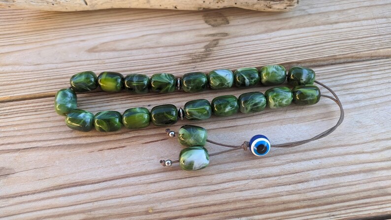 Op de afbeelding: Een groene en witte gebedskralenketting met een blauwe kwade oog charme. De kralen zijn gemaakt van een gemarmerd materiaal en zijn geregen aan een bruin koord.