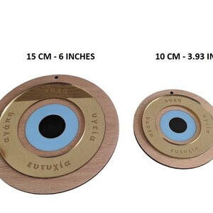 May include: Two round wooden ornaments with a blue evil eye design. The larger ornament is 6 inches in diameter and the smaller ornament is 3.93 inches in diameter. Both ornaments have gold lettering around the evil eye that reads "agapi", "tyhi", "ygeia", and "eytyhia".