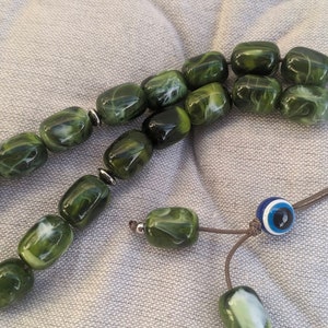 Op de afbeelding: Een groene en witte gebedsketting met een blauwe en witte kwade oog charme. De kralen zijn gemaakt van een gemarmerd materiaal en zijn geregen aan een bruin koord.