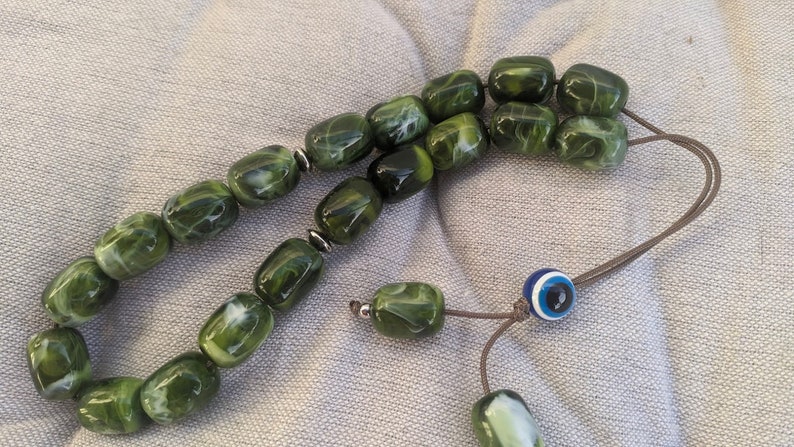 Op de afbeelding: Een groene en witte gebedsketting met een blauwe en witte kwade oog charme. De kralen zijn gemaakt van een gemarmerd materiaal en zijn geregen aan een bruin koord.