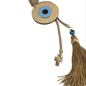 Op de afbeelding: Een gouden bedel met een blauw boze oog en de tekst "Yia sou Agia tou Xristou" hangend aan een beige touw met een gouden kruis en een gouden kwast.
