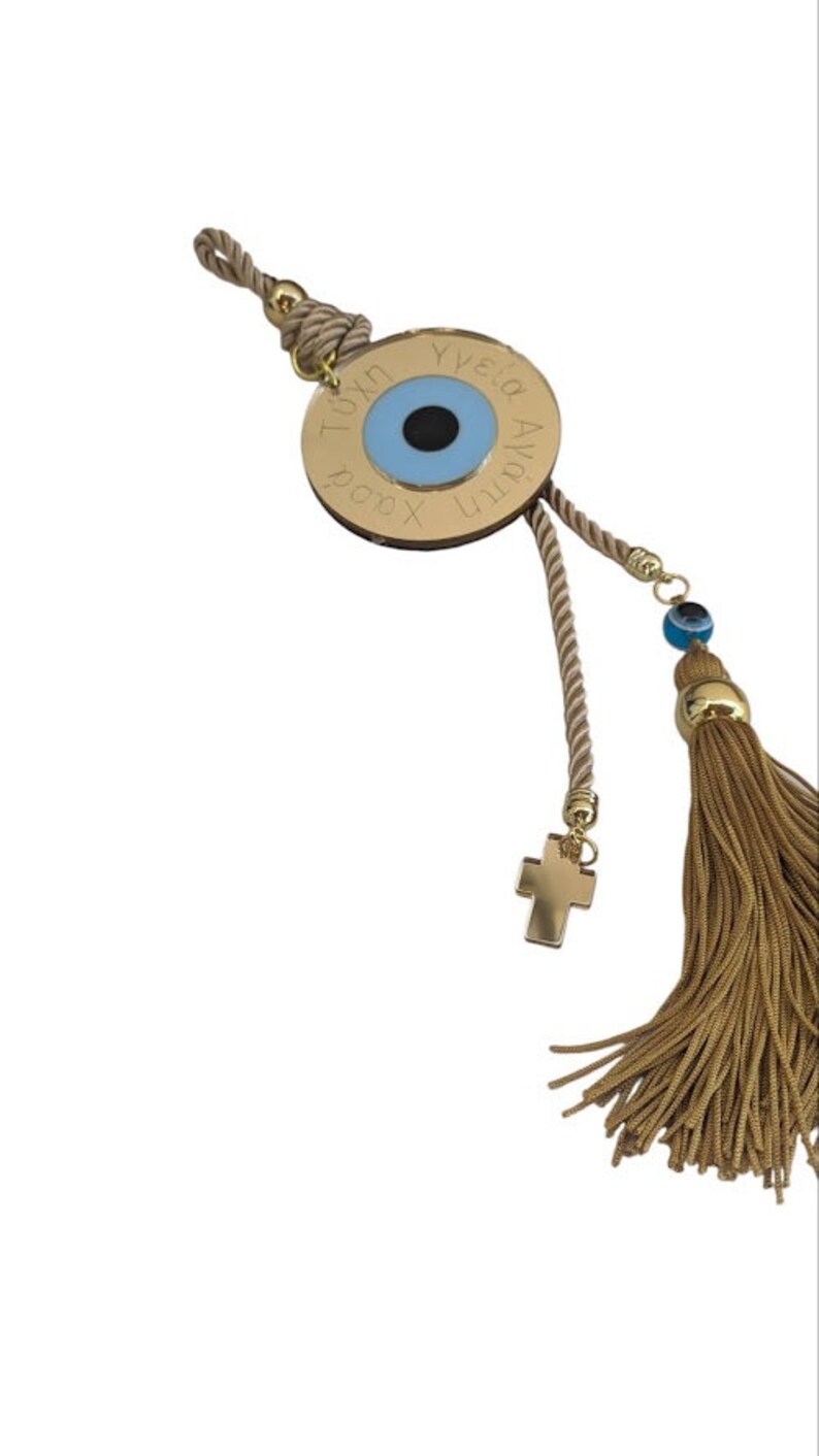 Op de afbeelding: Een gouden bedel met een blauw boze oog en de tekst "Yia sou Agia tou Xristou" hangend aan een beige touw met een gouden kruis en een gouden kwast.