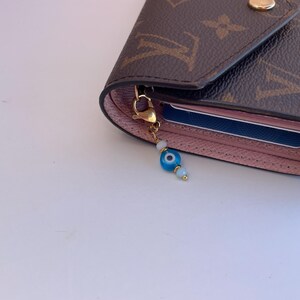 Pu&ograve; includere: Portafoglio marrone e rosa con chiusura dorata e un ciondolo a forma di occhio blu e bianco. Il portafoglio presenta un motivo monogram marrone e contiene una carta blu. Il ciondolo &egrave; fatto di perline blu e bianche.