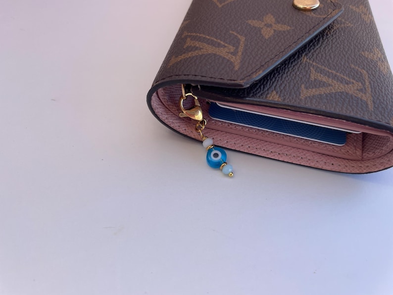 Pu&ograve; includere: Portafoglio marrone e rosa con chiusura dorata e un ciondolo a forma di occhio blu e bianco. Il portafoglio presenta un motivo monogram marrone e contiene una carta blu. Il ciondolo &egrave; fatto di perline blu e bianche.