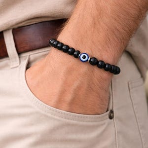 May include: Black beaded bracelet with an evil eye charm. The bracelet features small, round, black beads and a central charm with a blue and white eye design. The bracelet is worn on a wrist, with a neutral background.