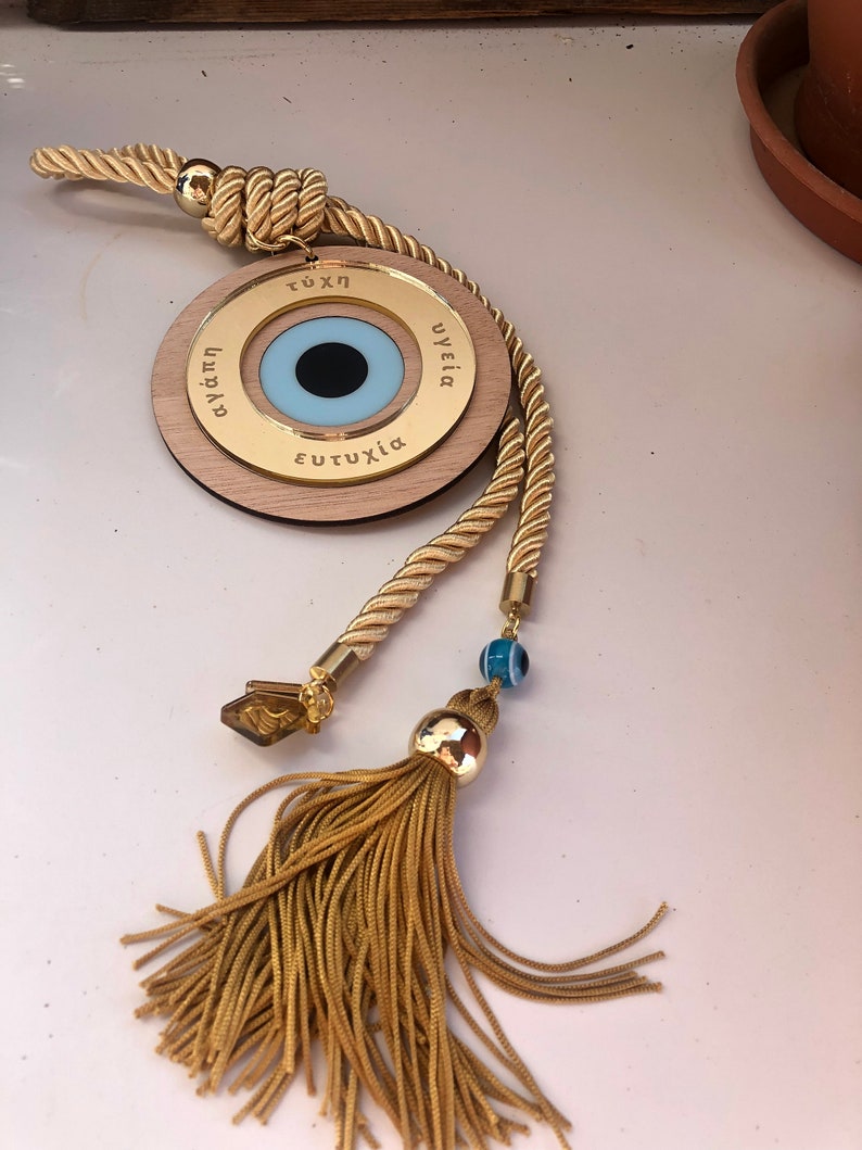 May include: A gold-colored wooden charm with a blue evil eye, a tassel, and a rope. The charm has the words "&epsilon;&upsilon;&tau;&upsilon;&chi;ί&alpha;", "&upsilon;&gamma;&epsilon;ί&alpha;", "&tau;ύ&chi;&eta;", and "&alpha;&gamma;ά&pi;&eta;" inscribed on it.