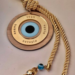 May include: A wooden circle with a blue evil eye charm and gold lettering that reads "&tau;ύ&chi;&eta;", "&upsilon;&gamma;&epsilon;ί&alpha;", and "&epsilon;&upsilon;&tau;&upsilon;&chi;ί&alpha;". The circle is attached to a gold rope with a tassel.