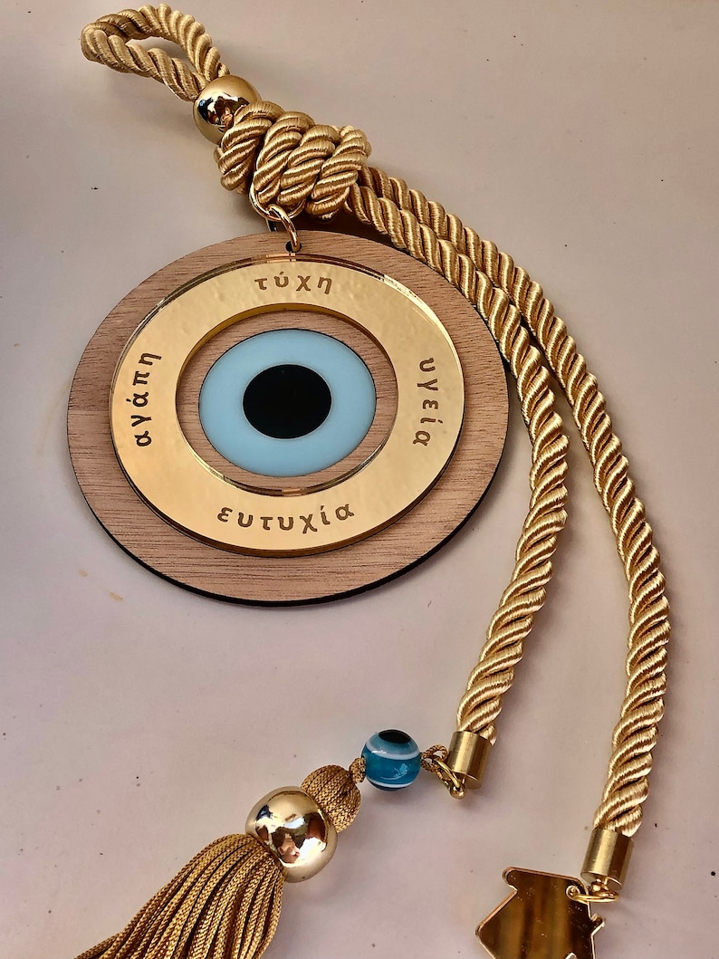 May include: A wooden circle with a blue evil eye charm and gold lettering that reads "&tau;ύ&chi;&eta;", "&upsilon;&gamma;&epsilon;ί&alpha;", and "&epsilon;&upsilon;&tau;&upsilon;&chi;ί&alpha;". The circle is attached to a gold rope with a tassel.