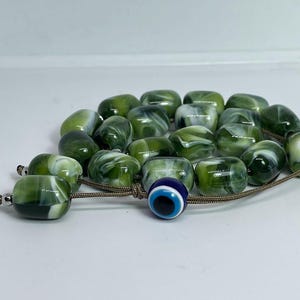 Op de afbeelding: Een groene en witte marmeren gebedsketting met een blauwe kwade oog-hanger. De kralen zijn geregen aan een bruin koord.
