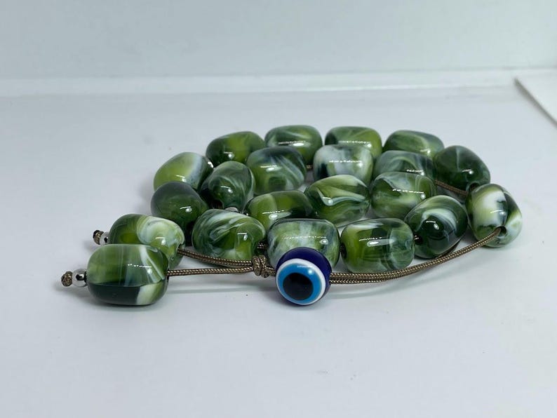 Op de afbeelding: Een groene en witte marmeren gebedsketting met een blauwe kwade oog-hanger. De kralen zijn geregen aan een bruin koord.
