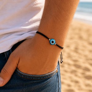 Peut inclure: Un bracelet en cordon noir doté d'une perle centrale ronde en verre avec un motif d'œil bleu, blanc et noir. Le bracelet est agrémenté de deux petites perles métalliques argentées de chaque côté du motif central et possède des extrémités réglables.