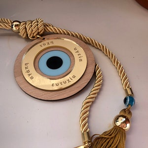 May include: A wooden charm with a blue evil eye and gold lettering around the edge. The charm is attached to a gold rope with a tassel and a blue evil eye bead.