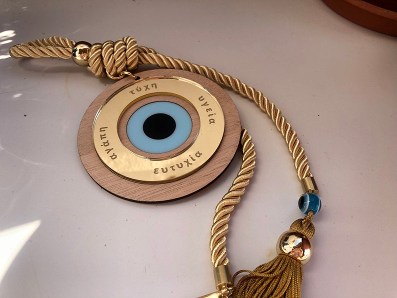 May include: A wooden charm with a blue evil eye and gold lettering around the edge. The charm is attached to a gold rope with a tassel and a blue evil eye bead.