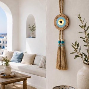 Op de afbeelding: Een ronde houten wandhanger met een centraal blauw en goudkleurig boze oog-ontwerp. Het hout is gegraveerd met de woorden Luck, Health, Love en Hope. Het hangt aan een dik touw van jute en is afgewerkt met twee lange beige kwastjes.