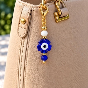 Peut inclure: Un sac à main beige texturé avec un fermoir en métal doré attaché à une breloque décorative. La breloque présente une perle en verre bleu en forme de fleur, une perle blanche, une petite perle bleue à facettes et des entretoises dorées.
