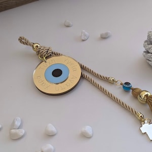 Op de afbeelding: Een ronde gouden hanger met een blauw boze oogontwerp en Griekse tekst. De hanger is bevestigd aan een beige kwast en een beige koord.