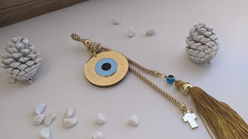 Op de afbeelding: Een ronde gouden hanger met een blauw boze oogontwerp en Griekse tekst. De hanger is bevestigd aan een beige kwast en een beige koord.