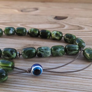Op de afbeelding: Groene en witte gebedskralen met een blauwe kwade oog charme. De kralen zijn gemaakt van een gemarmerd materiaal en zijn geregen aan een bruin koord.