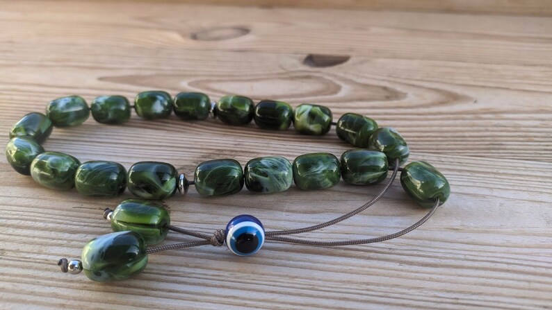 Op de afbeelding: Groene en witte gebedskralen met een blauwe kwade oog charme. De kralen zijn gemaakt van een gemarmerd materiaal en zijn geregen aan een bruin koord.