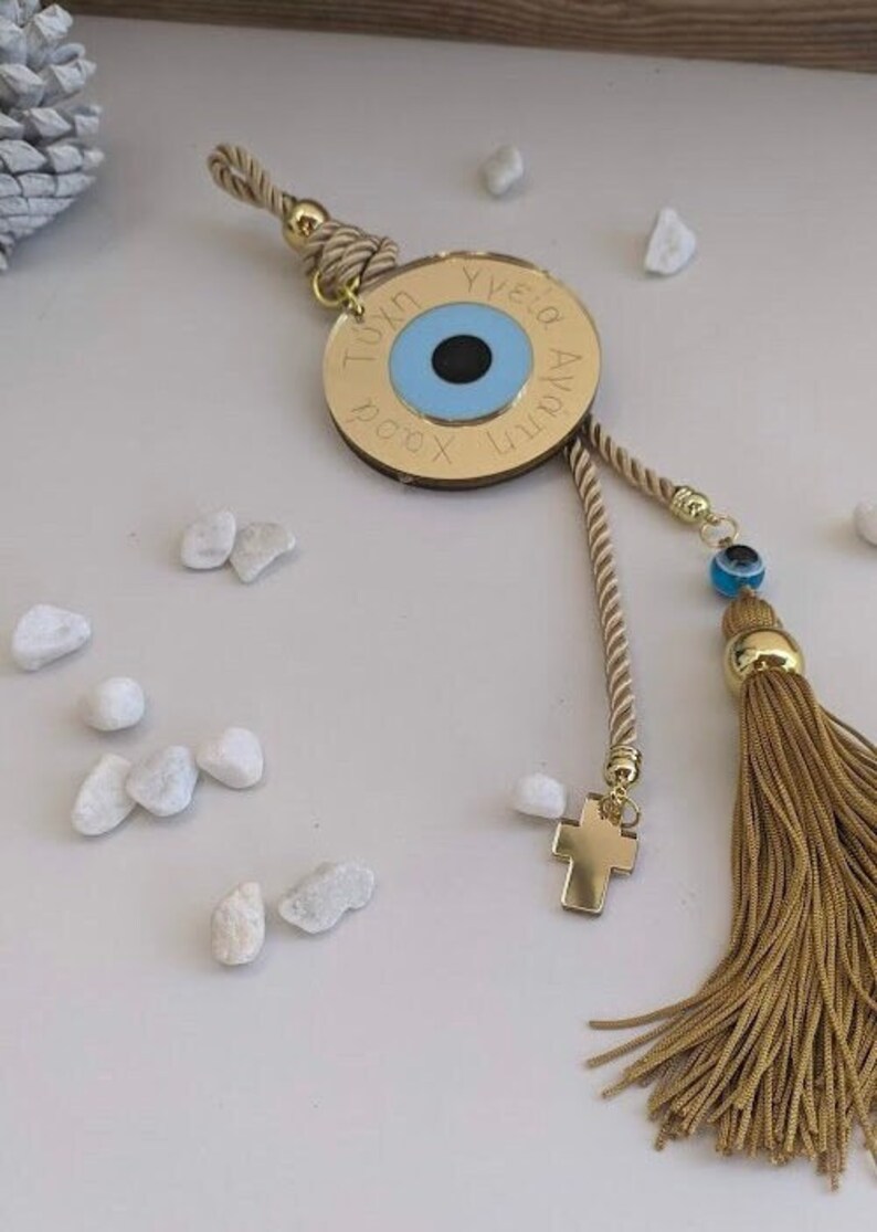 Op de afbeelding: Gouden bedel met een blauw boze oog, een gouden kruis en een gouden kwast. De bedel is gegraveerd met de Griekse woorden "Tuxh", "Ygeia", "Agapi" en "Xronia Polla".