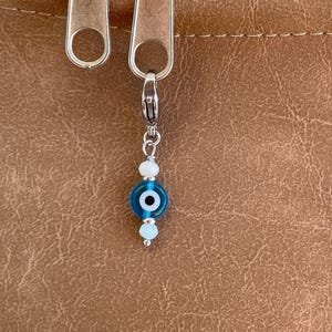 Pu&ograve; includere: Un ciondolo d'argento con una perla a forma di occhio blu e bianco, appeso a una cerniera. Il ciondolo ha una piccola perla blu rotonda con un centro bianco ed &egrave; accentuato da perline bianche pi&ugrave; piccole.