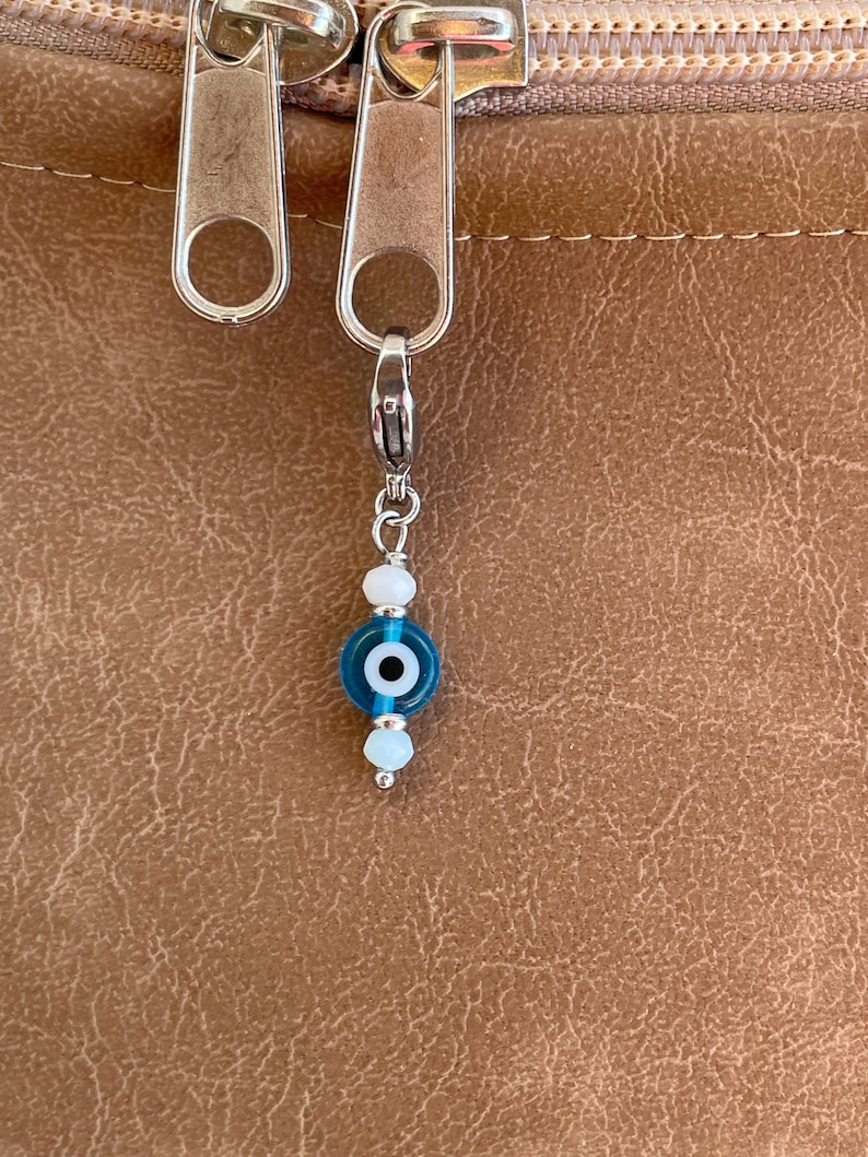 Pu&ograve; includere: Un ciondolo d'argento con una perla a forma di occhio blu e bianco, appeso a una cerniera. Il ciondolo ha una piccola perla blu rotonda con un centro bianco ed &egrave; accentuato da perline bianche pi&ugrave; piccole.