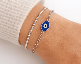 Può includere: Due braccialetti d'argento. Uno è in stile con perline, l'altro presenta un ciondolo a forma di occhio blu e bianco. Il ciondolo è di forma ovale con sfondo blu e centro bianco e nero. I braccialetti sono indossati su un polso.