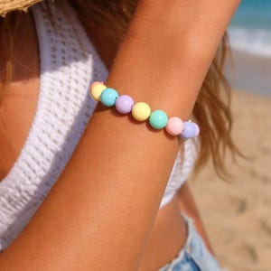 Könnte beinhalten: Ein Armband mit pastellfarbenen Perlen in Gelb, Blau, Lila und Rosa. Das Armband wird an einem Arm getragen, mit einem Strand und Meer im Hintergrund. Ein fröhliches, farbenfrohes Accessoire.