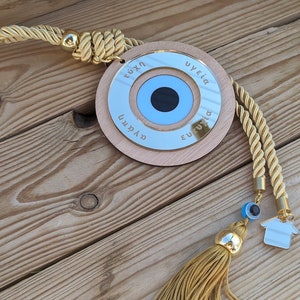 May include: A wooden circle with a blue evil eye charm and gold trim. The circle has the words "tyxh", "ygeia", "eytyxia", and "agaph" written around the edge. The circle is attached to a gold rope with a tassel.