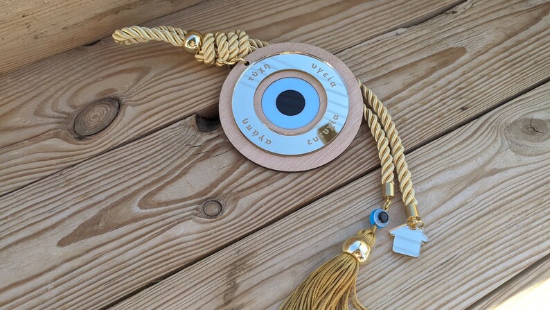 May include: A wooden circle with a blue evil eye charm and gold trim. The circle has the words "tyxh", "ygeia", "eytyxia", and "agaph" written around the edge. The circle is attached to a gold rope with a tassel.